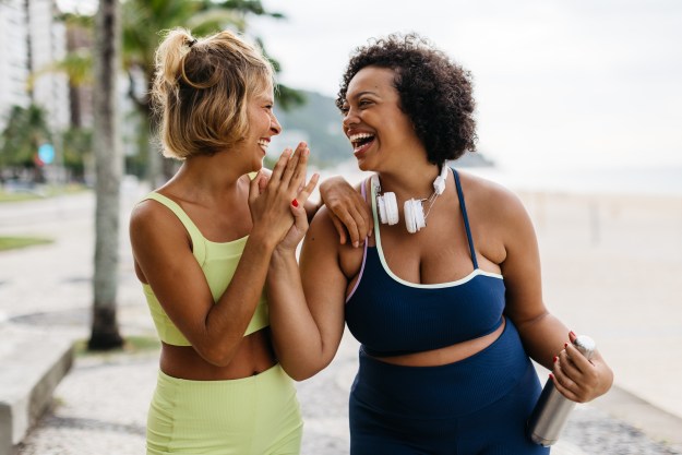 Two happy women in sportswear enjoy their workout routine on the ocean promenade, celebrating with a high-five. Female friends with different body shapes embracing a lifestyle of fitness.